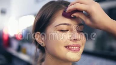不可识别的视觉应用液体基础`年轻妇女的脸使用海绵。 美容院的专业化妆